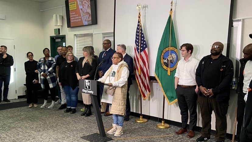 A woman stands at a podium during a press event. Fifteen people stand behind her.
